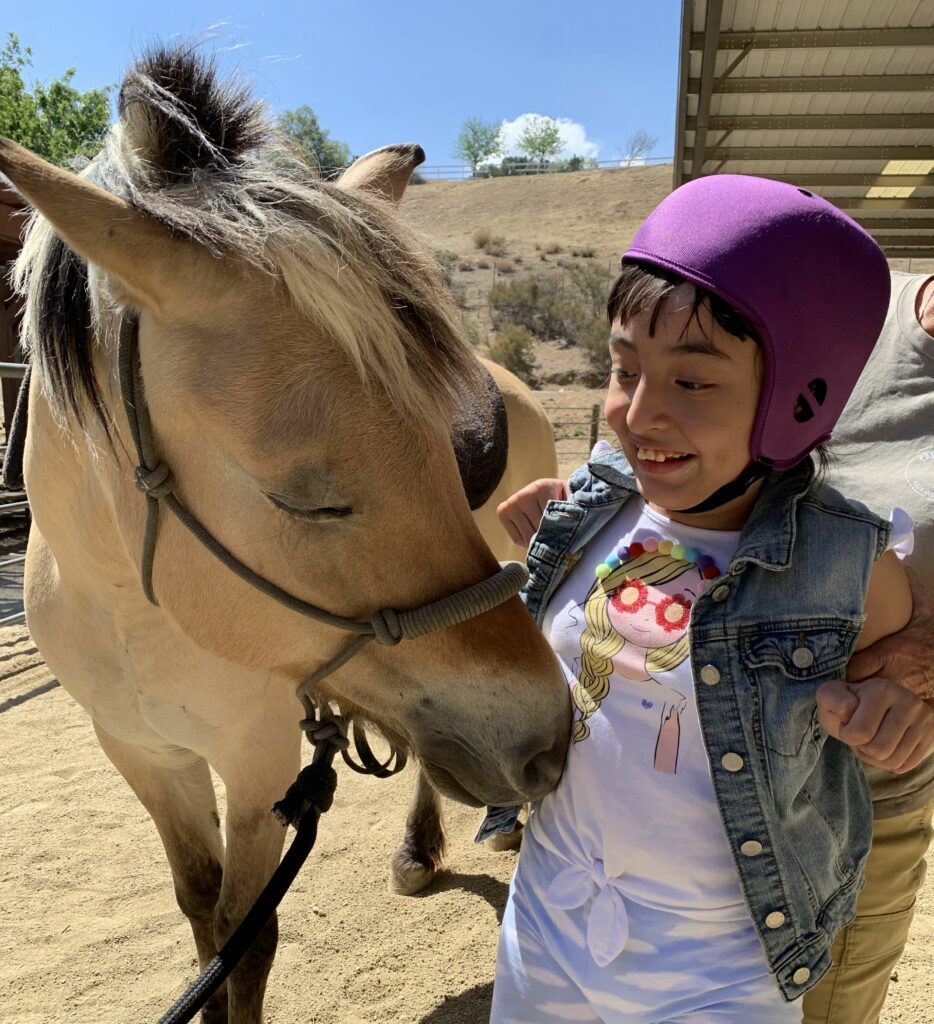 Young girl wearing a helmet smiling and petting a horse during the day